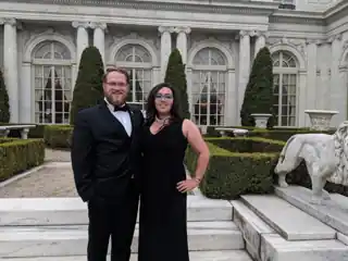 Laura and Bryce in formal clothes beside an ornate garden and building.