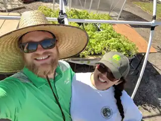 Laura and Bryce in a greenhouse, wearing farm hats.