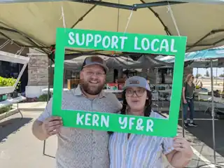 Laura and Bryce holding a green Support Local Kern YF&R frame at an outdoor market.